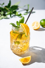 Fototapeta premium Lemonade drink or cold tea with fresh lemon, mint and ice in a jar. Refreshing citrus mojito cocktail on a light background with fresh fruits and shadows. Homemade healthy and detox drink concept