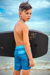 Cute child surfer boy standing wet on the beach holding his boogie board under his arm