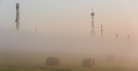 Rural scene Romania. Foggy rural landscape in the morning with cell phone tower and electricity transmission pylon,  straw bales in front of it. - 648963231