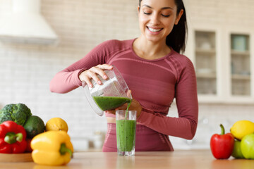 Woman In Fitwear Pouring Green Smoothie In Glass At Kitchen