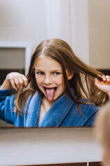 Beautiful funny red-haired smiling teen girl in a bathrobe, the child grimaces, plays with hair, showing tongue, looking in the mirror in the bathroom in the morning. Photography, close-up portrait.