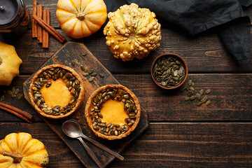 Obraz na płótnie Canvas Two mini pumpkin tarts decorated with roasted pumpkin seeds on rustic wooden board with various colorful decorative pumpkins, cinnamon sticks, dark napkin and candle. Overhead view, copy space