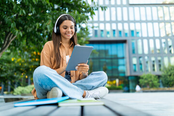 Cool young woman student sitting outdoors, using digital tablet, headphones