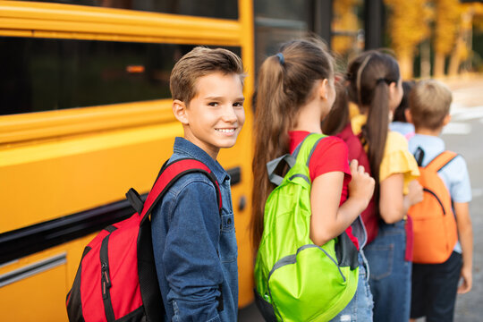 Portrait Of Smiling Boy With Backpack Boarding Yellow School Bus With Classmates