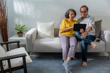 Senior Asian couple sitting happily relaxing on the sofa, using tablet devices with hands, smiling and laughing together.