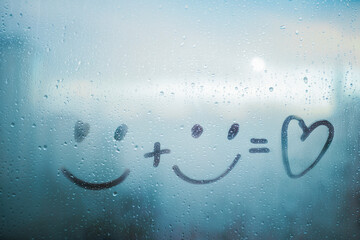 Two drawn smile face and doodle heart on blue foggy glass of window, concept positive photo