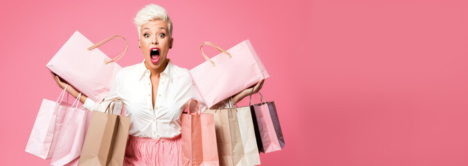 A middle-aged hipster woman holding shopping bags, surprised face with mouth open, pastel pink background copyspace, low prices sale and discounts for black friday concept
