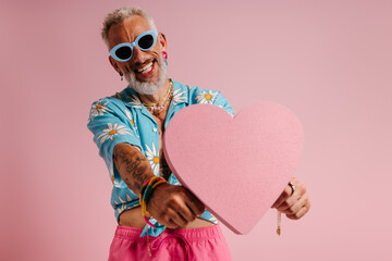 Happy mature gay man in stylish glasses holding heart shape and smiling against pink background