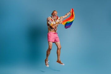 Full length of mature gay man carrying rainbow flag and smiling while jumping against blue background