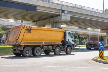 Yellow dump truck with a scratched body drives along a city street under a bridge road.