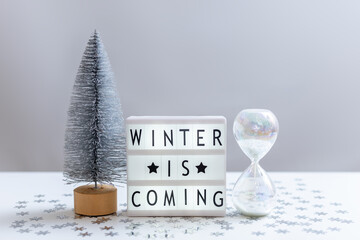 Still life with a silver Christmas tree, a glass hourglass and a lightbox with the inscription Winter is coming
