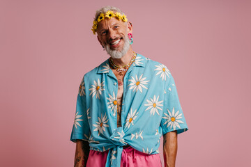 Obraz premium Happy senior man in stylish shirt and flower wreath on head standing against pink background