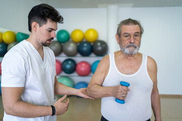 Calm doctor helping senior patient with dumbbells in clinic