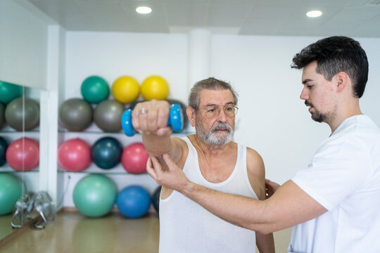 Calm doctor helping senior patient with dumbbells in clinic