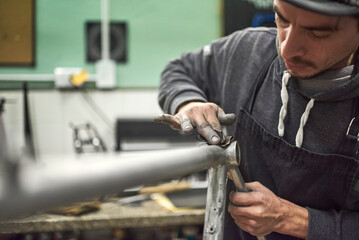 Latin man sanding an unpainted bicycle frame as part of the process of a bike renovation work made at his workshop. Selective focus composition.
