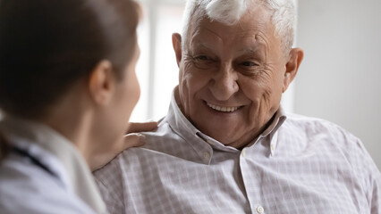 Close up of optimistic old 80s man talk joke with caring female nurse in retirement house, smiling...