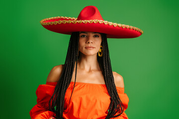 Gorgeous young Mexican woman in Sombrero looking at camera while standing against green background