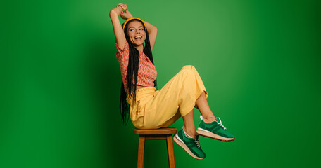 Beautiful young woman with dreadlocs looking happy while sitting on the chair against green...