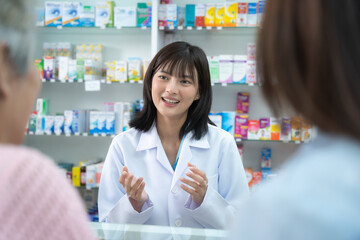 Young Asian pharmacist talking with customer at pharmacy counter. She tells customers about drug...