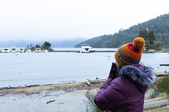 Woman dressed in winter outfit looking towards the lake with fog and with some boats.