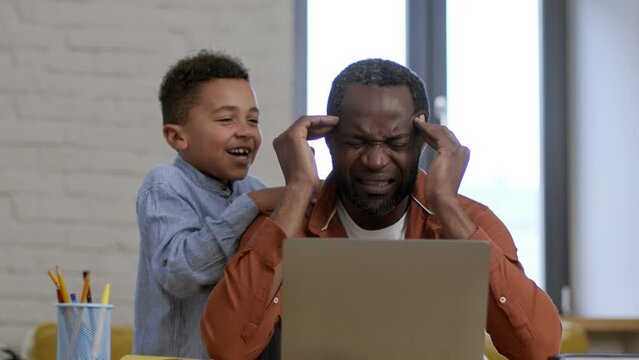 Unhappy african american man suffering from acute headache at home office, active annoying son shouting at shaking him