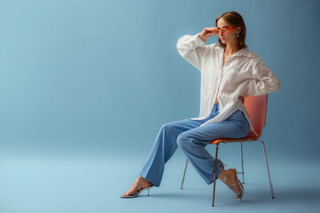  Fashionable young woman wearing trendy orange sunglasses, white linen shirt, trousers, metallic strap sandals, posing on blue background. Full-length studio portrait. Copy, empty space for text