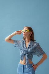 Fashionable young woman wearing trendy yellow sunglasses, blue chiffon knotted blouse, posing on blue background. Studio portrait. Copy, empty space for text