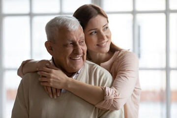 Smiling loving millennial girl child hug elderly 80s dad look in distance dreaming at home together, happy senior father and grownup adult daughter embrace feel optimistic imagine good future