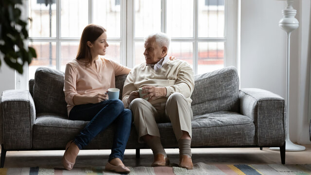 Happy elderly father sit on couch in living room talk with adult daughter drinking tea together, smiling senior dad and adult millennial girl enjoy family time weekend in retirement house or home