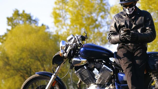male biker motorcyclist in a leather jacket and helmet enjoys a smartphone while standing by a chopper motorcycle in the fall. Mobile application for motorcyclists.