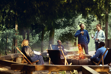 Multicultural group of friends enjoying beer by the campfire on a summer evening. Camping, tourism, holiday, outdoors concept. Barbeque party. Corporate team building event. Active lifestyle. - obrazy, fototapety, plakaty