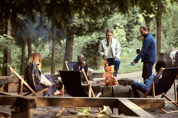 Multiethnic group of friends hanging out by the campfire and drinking beer. A man pours lighter...
