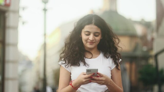 Portrait of young curly woman hold smartphone scrolling social media texting browsing online while walking at urban city Pretty female relax enjoying great day and looking at screen at street outdoors