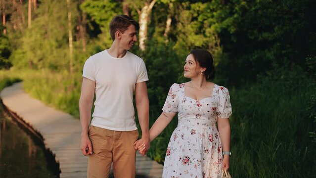 A guy and a girl walk along a wooden path by the water holding hands. Lovers look at each other and smile