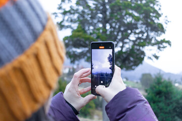 Woman recording video through her smartphone in wooded and cloudy landscape.