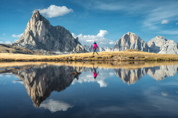 Walking woman and mountains reflected in lake at sunset in autumn in Dolomites, Italy. Standing girl on the shore of lake is looking on high rocks and blue sky with clouds in fall. Reflection in water - obrazy, fototapety, plakaty