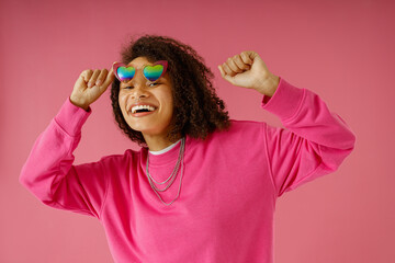 Joyful woman with curly hair is touching heart-shaped glasses on pink studio background