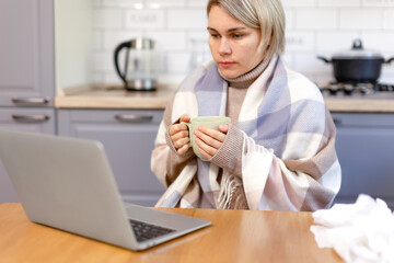 A woman wrapped in a scarf works on a laptop and warms herself with hot tea. A woman works at home...