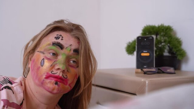 Smartphone alarm clock rings on bedside table, swollen disheveled woman wakes up, raises her head, smeared cosmetics, face painted with markers Hangover, morning after stormy party, bachelorette