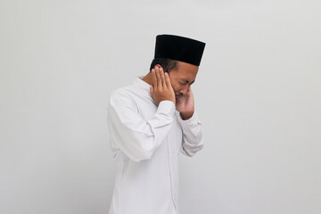 Young Indonesian man, wearing a songkok, peci, or kopiah, holds his hand near his ear, suffering...