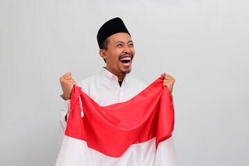 an Excited young Indonesian man, wearing a songkok, peci, or kopiah, is holding an Indonesian flag to celebrate Indonesia Independence Day on 17 August, isolated on white background