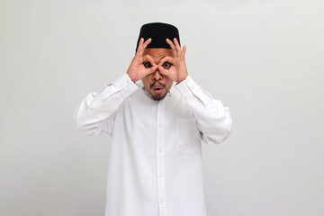 Obraz premium Excited young Indonesian man, wearing a songkok, peci, or kopiah, makes the OK sign gesture with his fingers, acting as binoculars with his eyes peeking through them, isolated on white background