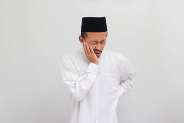 Unhappy young Indonesian man, wearing a songkok, peci, or kopiah, holding his cheek with his hand and showing a painful expression due to toothache or dental illness, isolated on white background