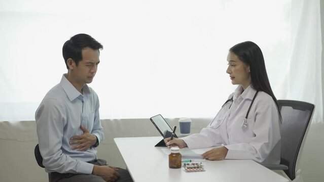 Young Asian doctor in white medical uniform uses clipboard to discuss results or symptoms with male patient sitting at table in clinic to discuss health issues.