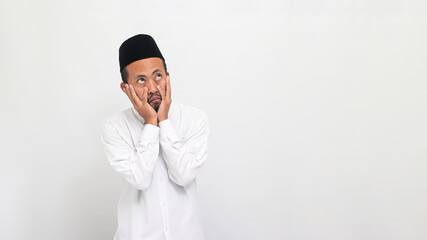 A funny young Indonesian man wearing a songkok or peci or kopiah, keeps his hand on his cheeks while looking up at empty copy space, looking bored, isolated on a white background