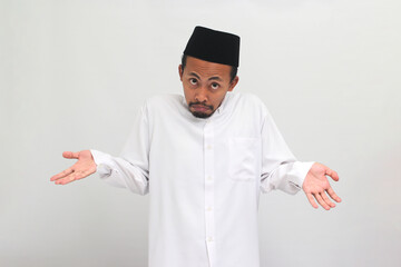 Confused young Indonesian man, wearing a songkok, peci, or kopiah, is shrugging his shoulders and looking at the camera with a puzzled expression, isolated on a white background