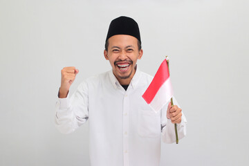 An excited young Indonesian man, wearing a songkok, peci, or kopiah, is celebrating Indonesian Independence Day on 17 August by holding a small Indonesian flag, isolated on white background