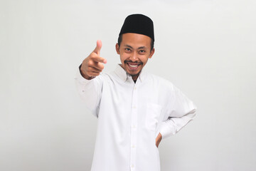 Confident young Indonesian man wearing a songkok or peci or kopiah, is pointing his finger at the camera and smiling, choosing, inviting people, or recruiting, isolated on a white background