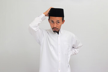 Pensive young Indonesian man, wearing songkok, peci, or kopiah, is scratching his head while looking at the camera, deep in thought, trying to find a solution, isolated on white background