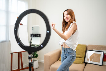 Young trendy influencer asian woman dancing on mobile phone at home in living room. Creator vlogger talent dancing enjoy hobby content recording show video sharing on social media.